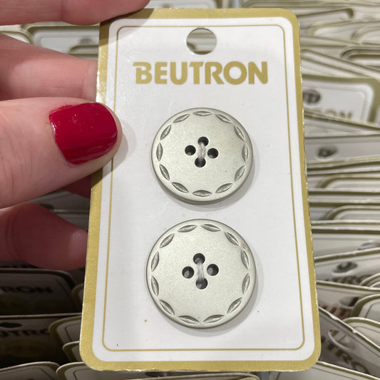Two decorative buttons on a Beutron branded card held by a hand with red nail polish.