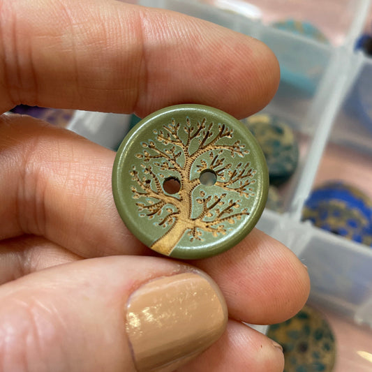 Hand holding a decorative button with intricate design, with a container of similar buttons in the background.
