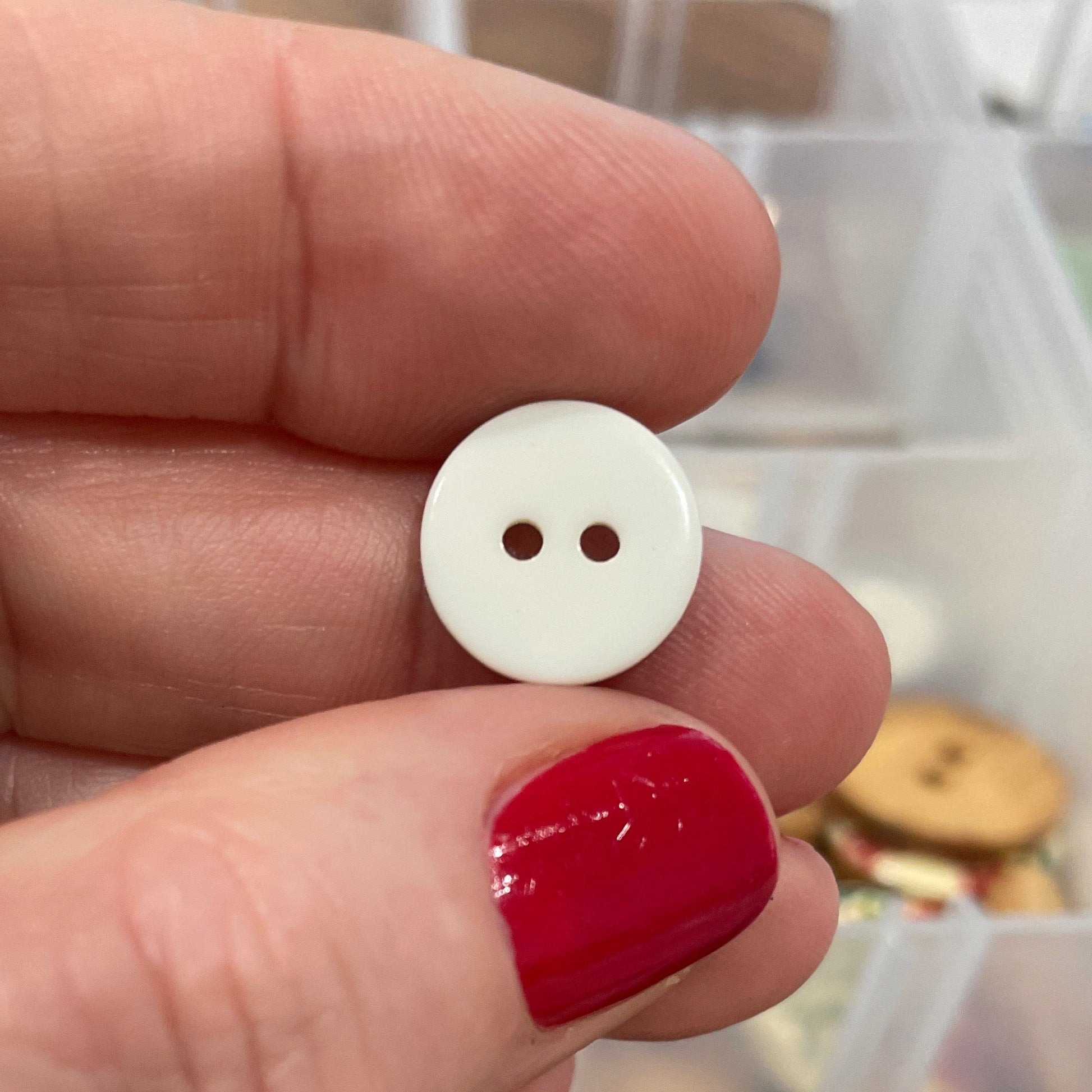 White button held by a hand with red nail polish, against a background of button assortment.