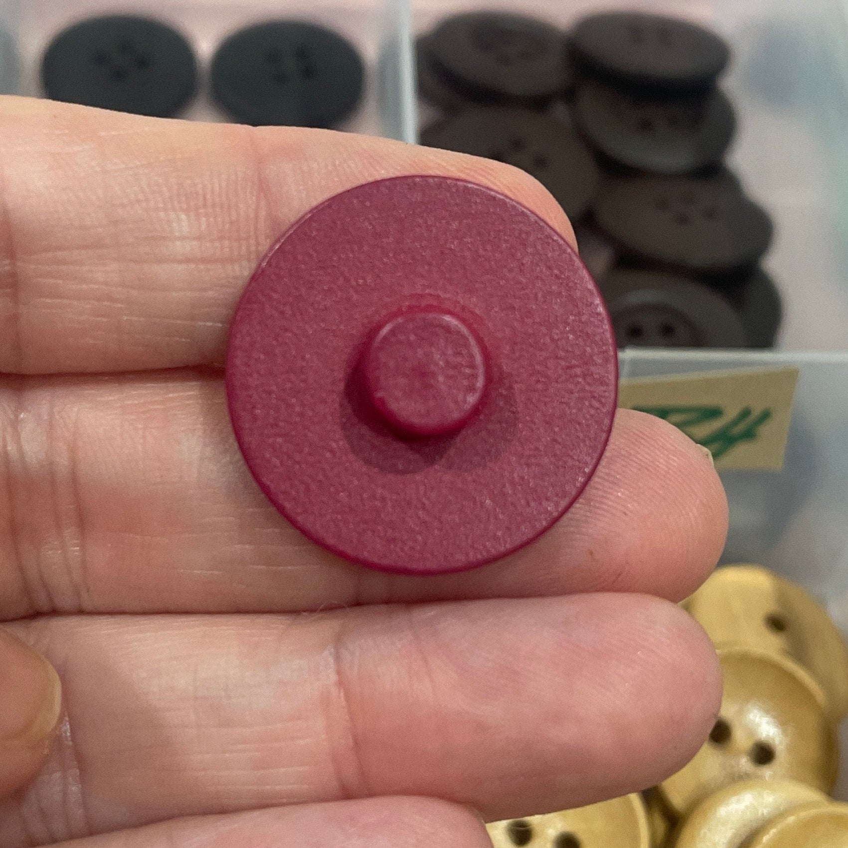 Hand holding a pink button with a container of buttons in the background