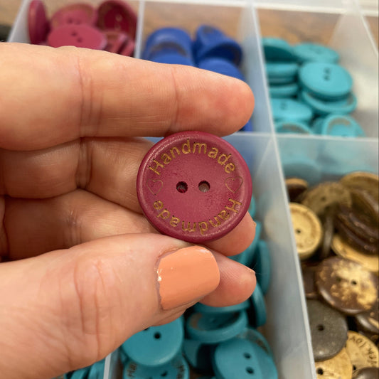 Hand holding a red button with 'Handmade' text, surrounded by various buttons in a container.