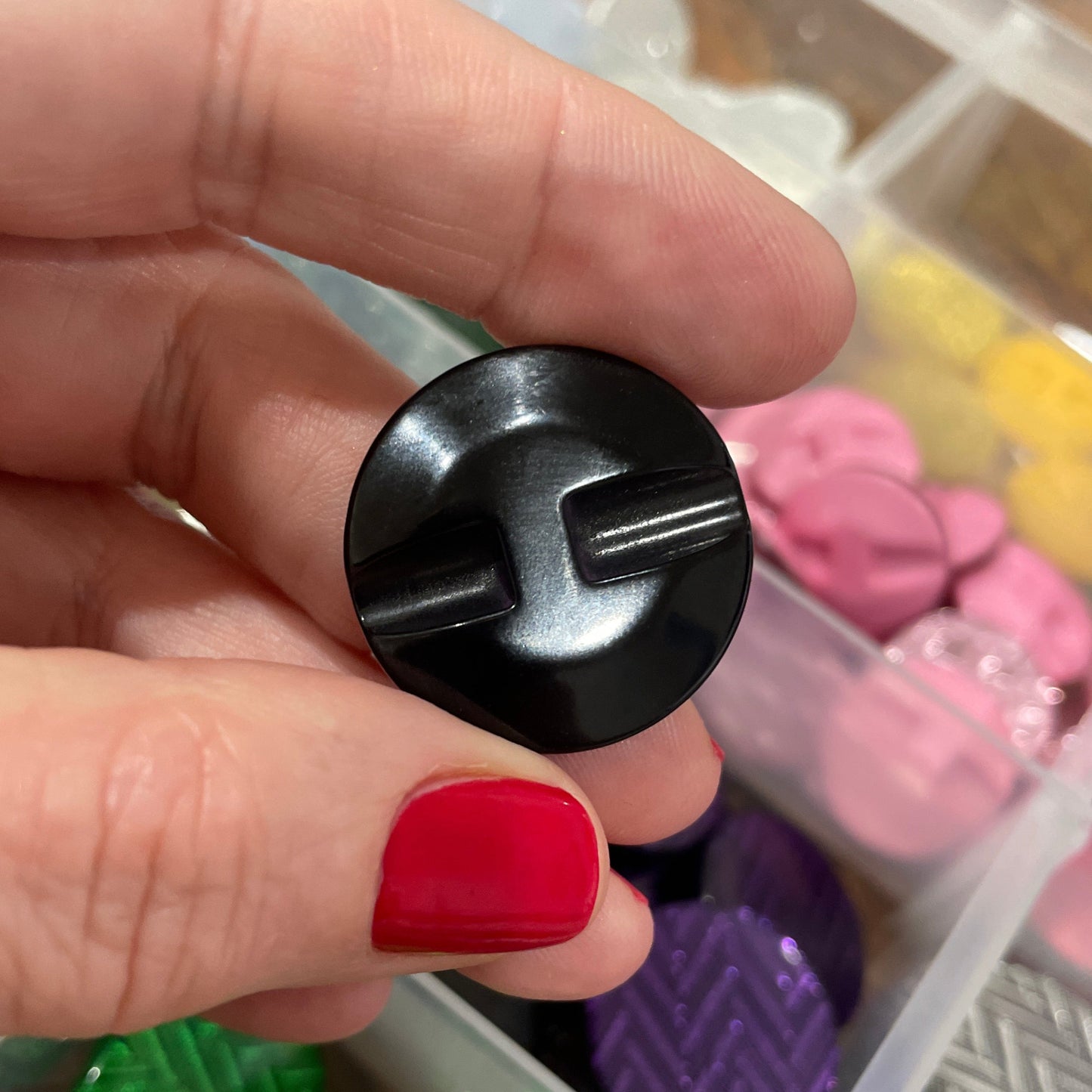 Black button held by a hand with red nail polish, against a background of colorful buttons in a container.