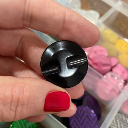 Black button held by a hand with red nail polish, against a background of colorful buttons in a container.