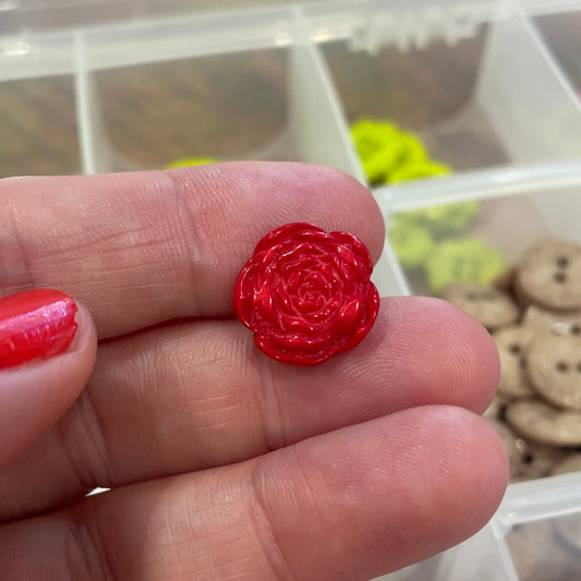 Red rose-shaped object on a hand with a blurred background of a storage box with buttons and other items.