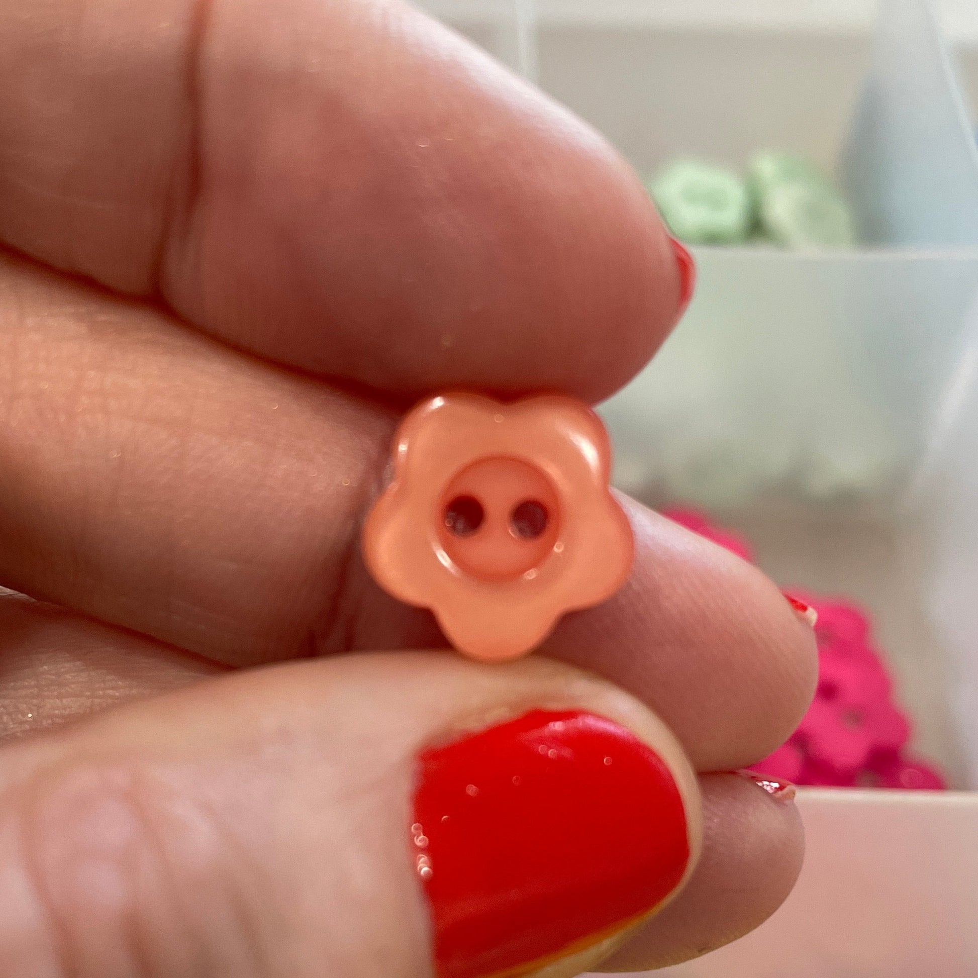 Hand holding two small flower-shaped buttons with a container of more buttons in the background.