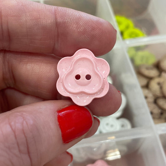 Pink flower-shaped buttons held between fingers with a container of similar buttons in the background.