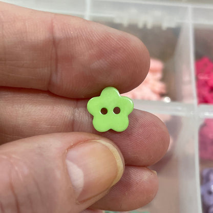 Hand holding a green flower-shaped button with a container of assorted buttons in the background.
