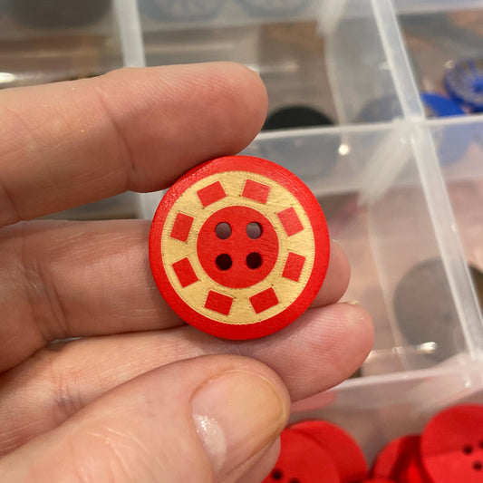 Red button with four holes held by a hand, with a container of buttons in the background.