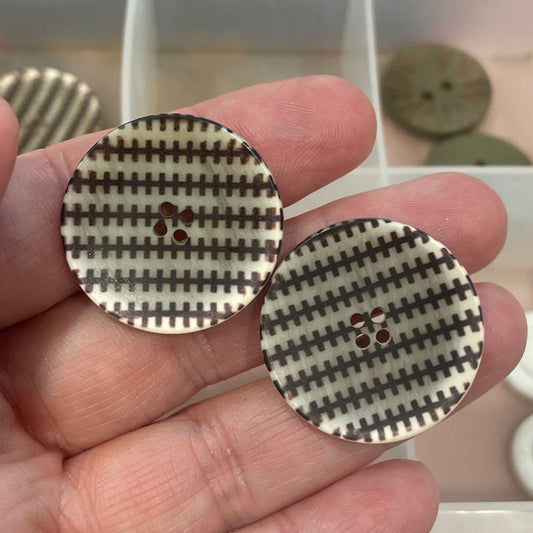 Hand holding two round buttons with a checkered pattern in front of a clear container with more buttons.