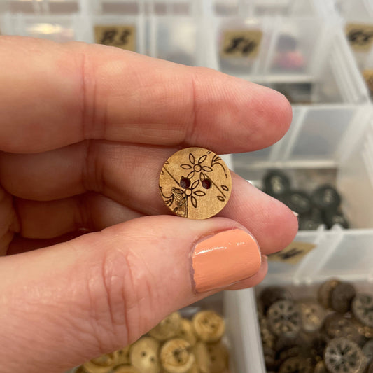 Hand holding a small wooden button with intricate design against a background of similar buttons in a container.