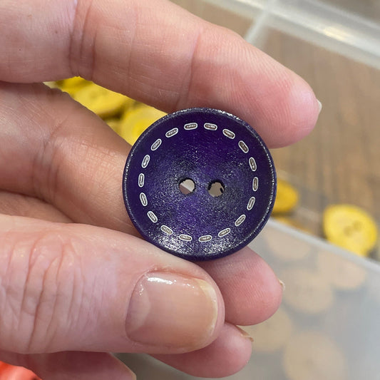 Hand holding a purple button with a white center, surrounded by various buttons in a container.
