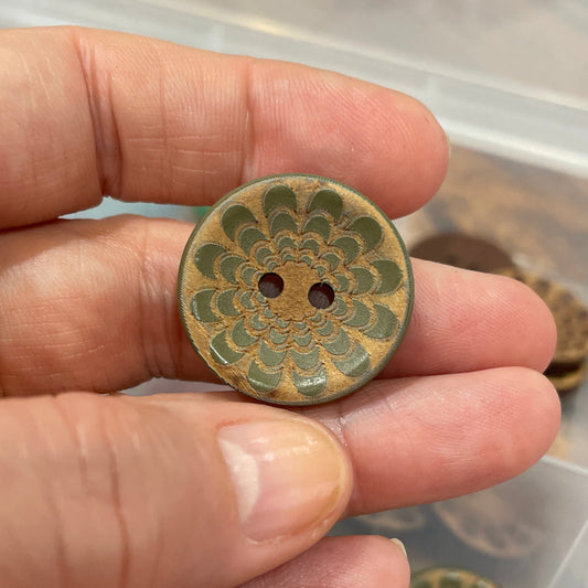 Hand holding a decorative button with a detailed design, against a background of similar buttons in a container.
