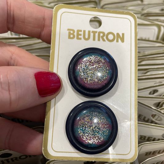 Pair of glittery buttons on a BEUTRON card held by a hand with red nail polish.