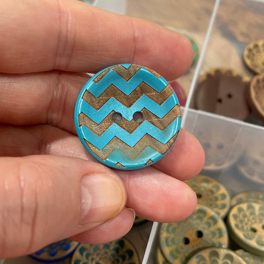 Hand holding a blue and gold zigzag patterned button with a container of similar buttons in the background.