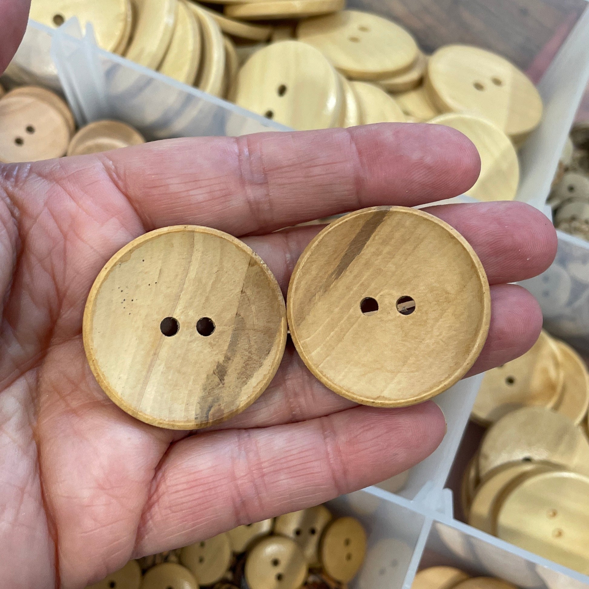 Hand holding two wooden buttons with a background of more buttons in a container.