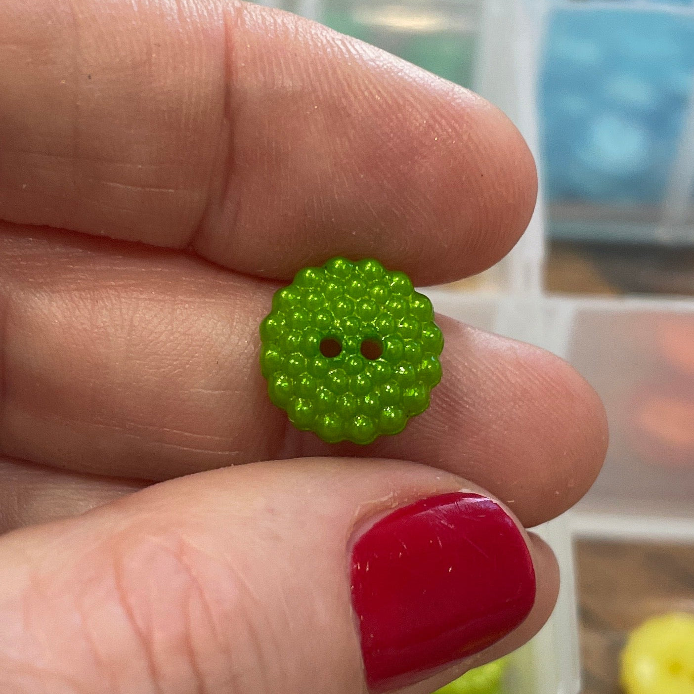 Green button held between fingers with a container of buttons in the background