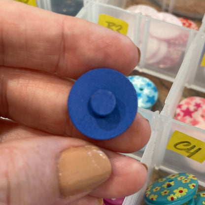 Hand holding a blue button with a container of assorted buttons in the background
