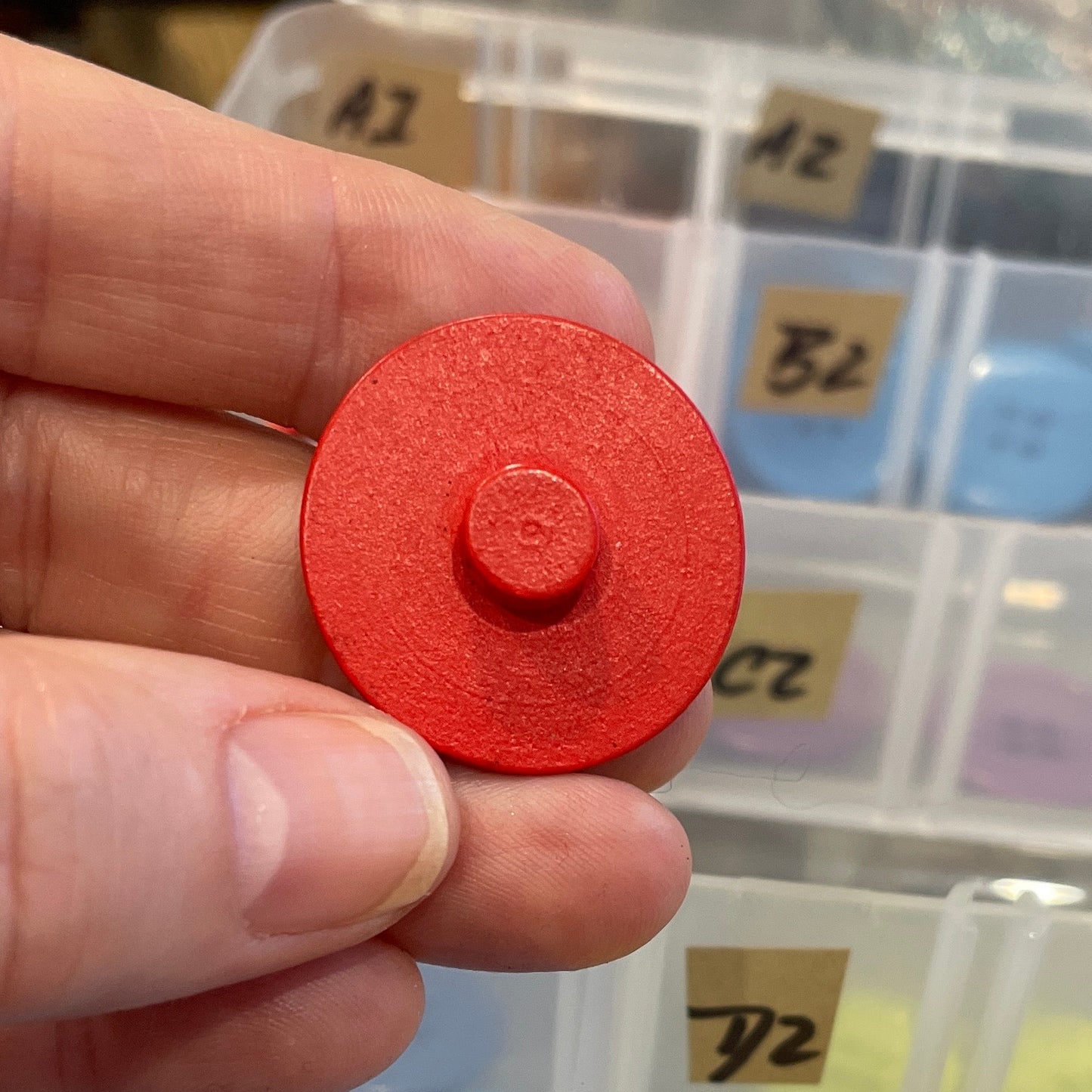 Hand holding a red button with a container of assorted buttons in the background