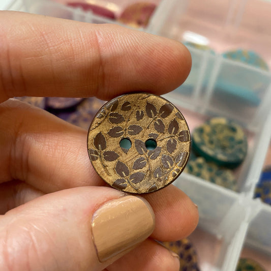 Hand holding a floral-patterned button with a container of buttons in the background