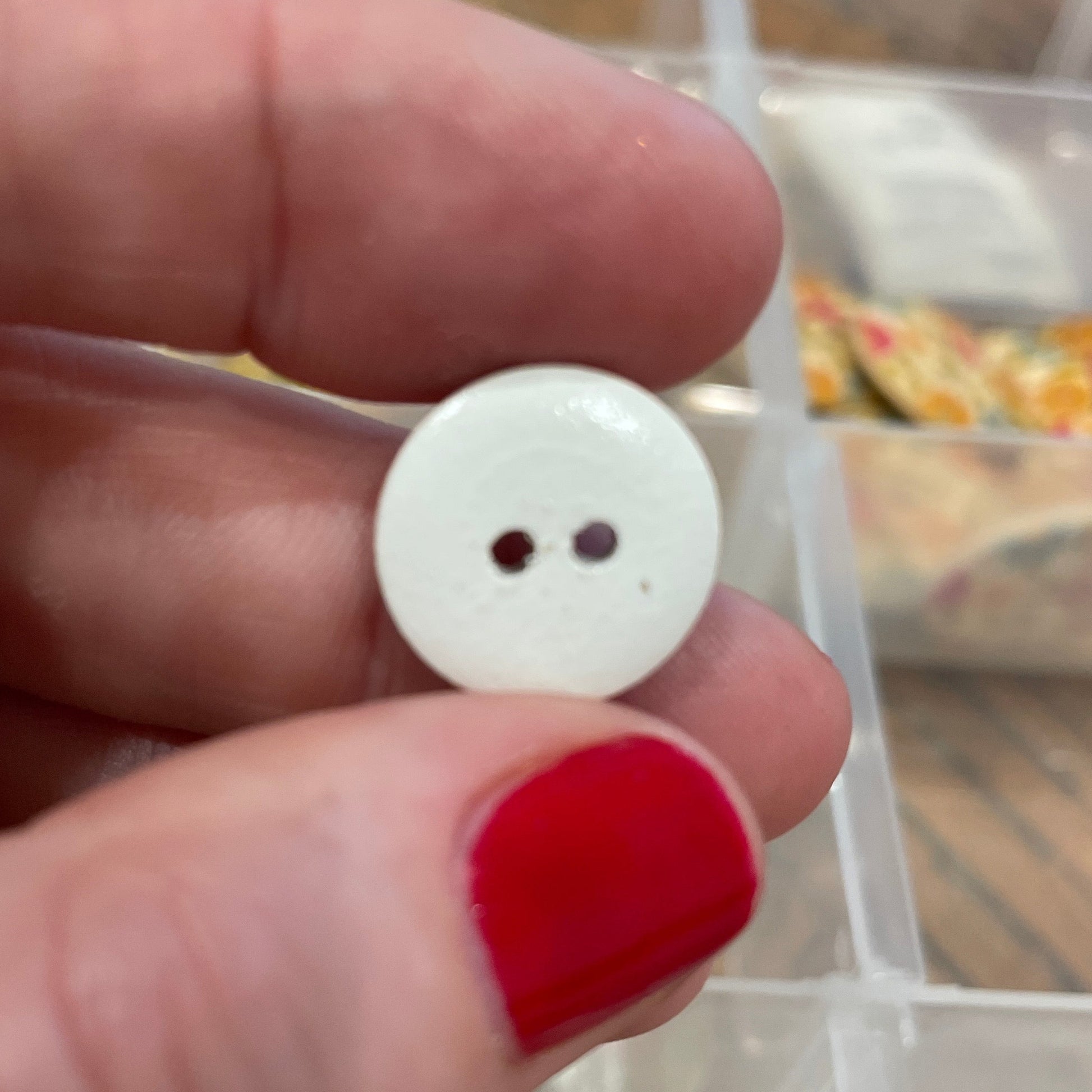 Hand holding a white button with two holes, with a background of various buttons in a container.