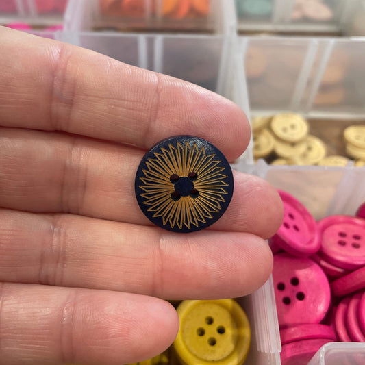Black button with gold design held between fingers, with a container of colorful buttons in the background.