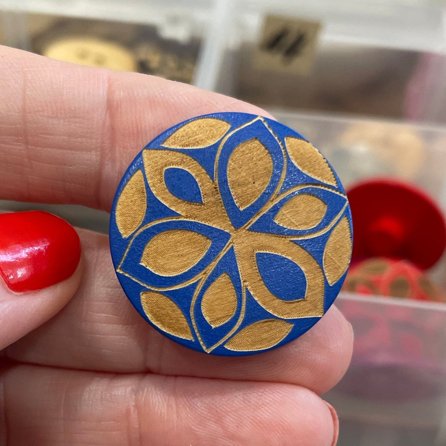 Hand holding a blue decorative button with a background of assorted buttons in clear containers.