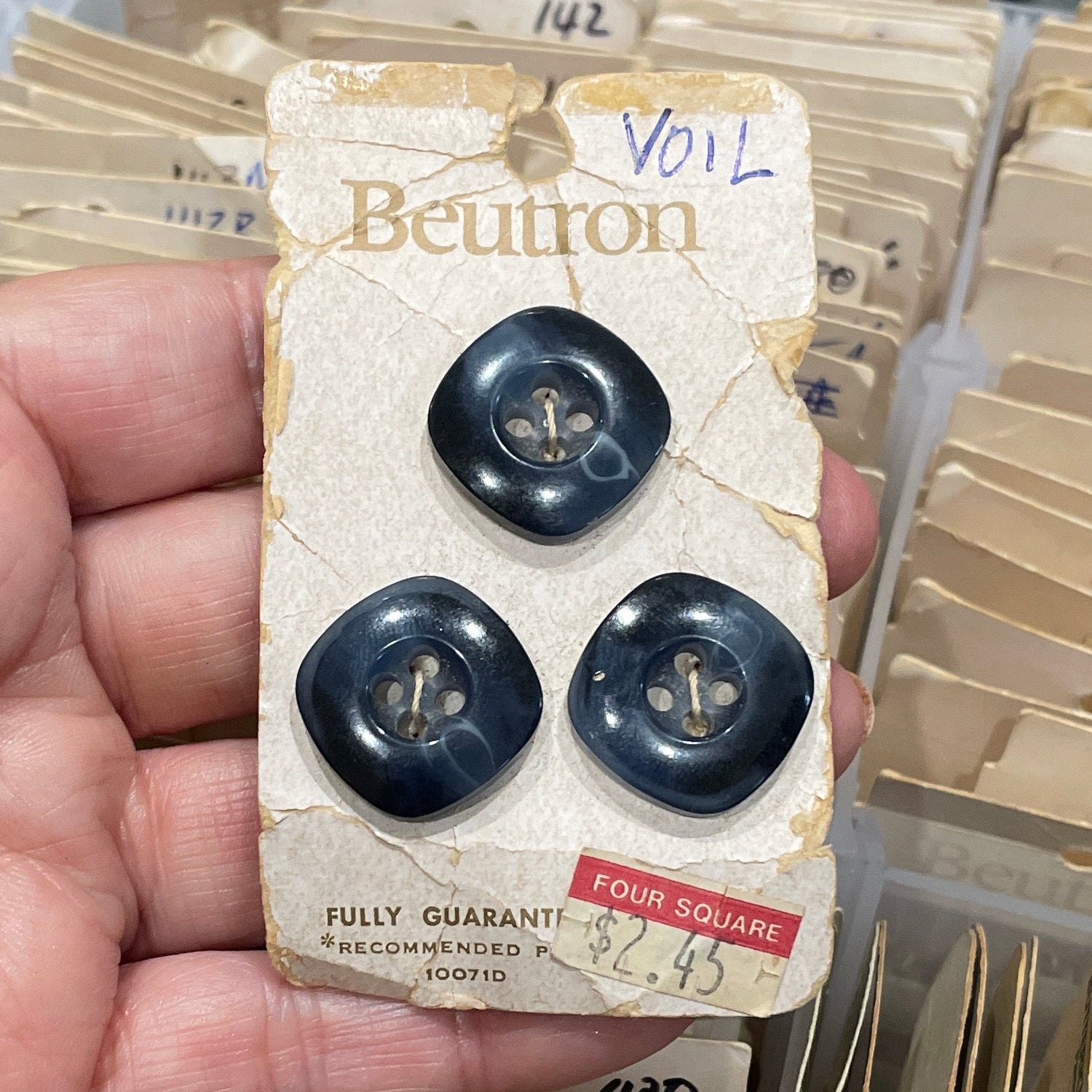 Hand holding a package of black buttons with a visible brand label, against a background of stacked similar packages.