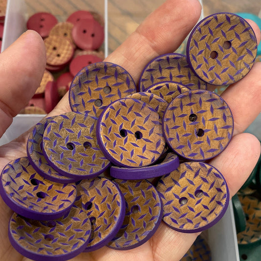 Hand holding purple and gold patterned buttons with a container of buttons in the background.