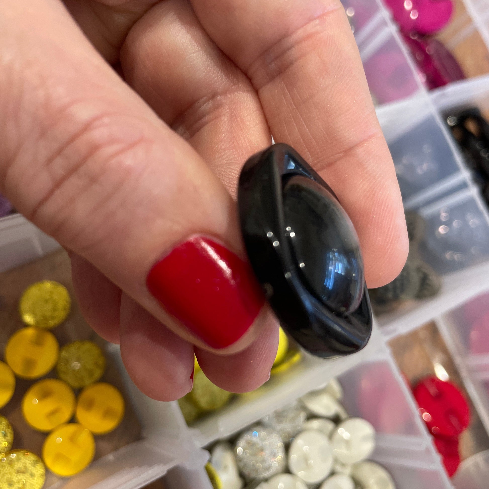 Black button held by a hand with red nail polish, surrounded by colorful buttons in the background.