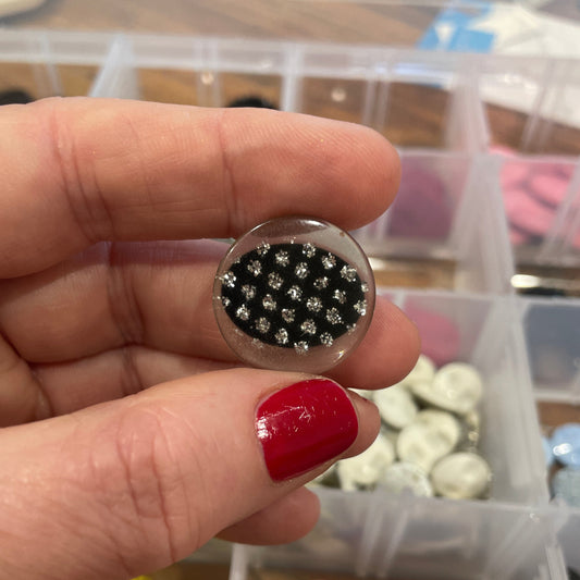 Close-up of a hand holding a small round object with black and silver pattern, against a background of colorful buttons.