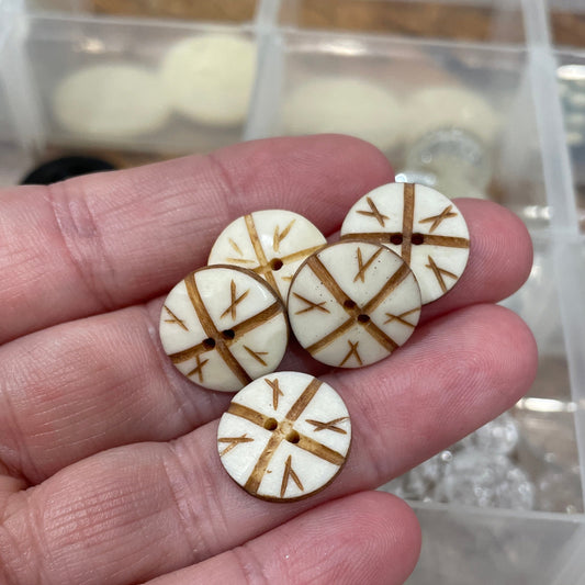 Hand holding four round wooden buttons with gold patterns against a blurred background of more buttons.