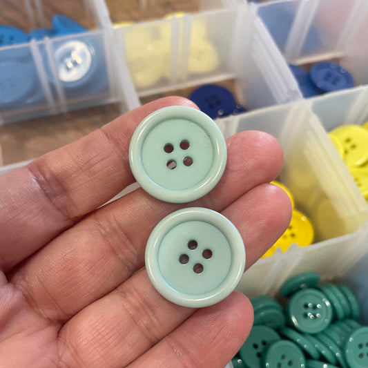 Two light green buttons held between fingers with a container of assorted buttons in the background.