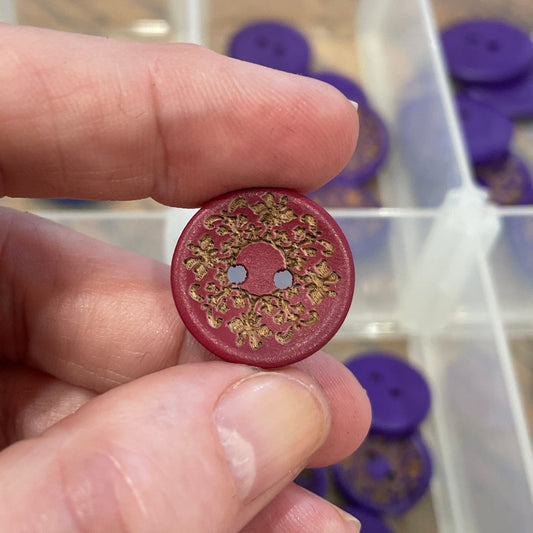 Hand holding a decorative red button with gold patterns, with a container of similar buttons in the background.