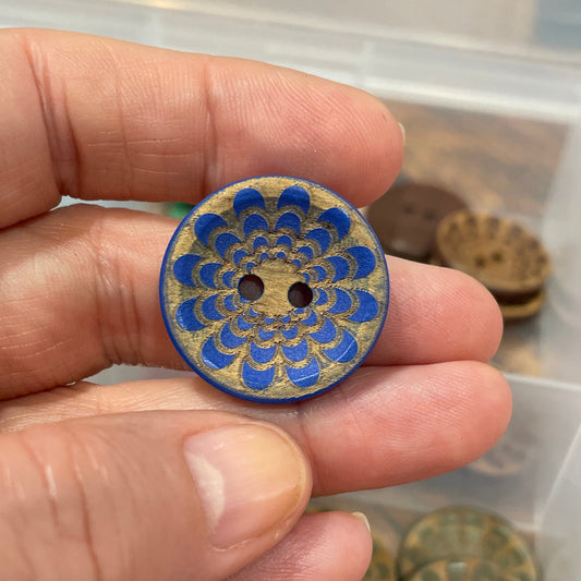 Hand holding a decorative button with intricate design, set against a background of similar buttons in a box.