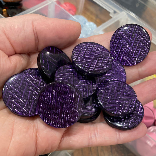Hand holding purple circular objects with a container of assorted colorful objects in the background.