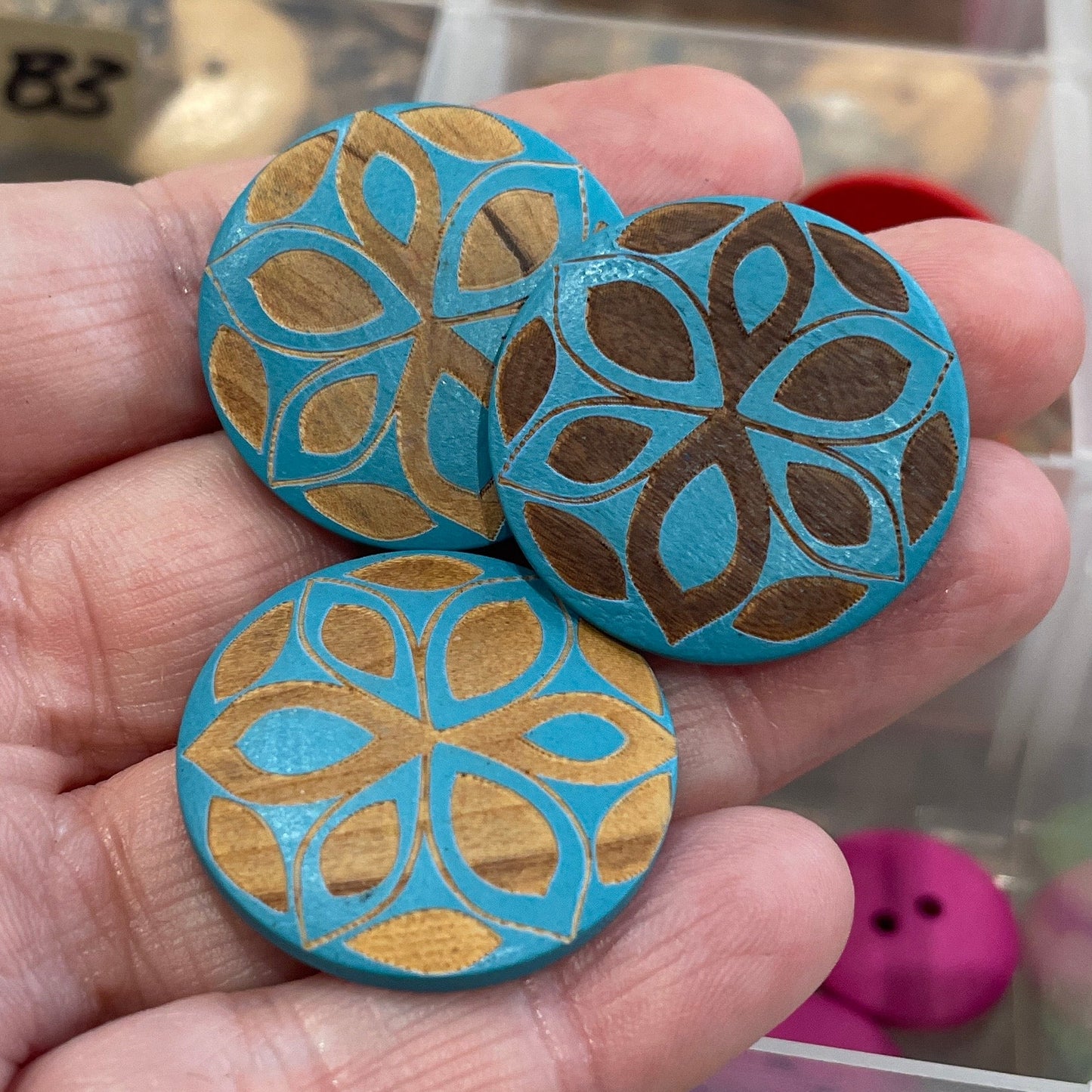 Hand holding decorative buttons with a patterned design in front of a container with more buttons.