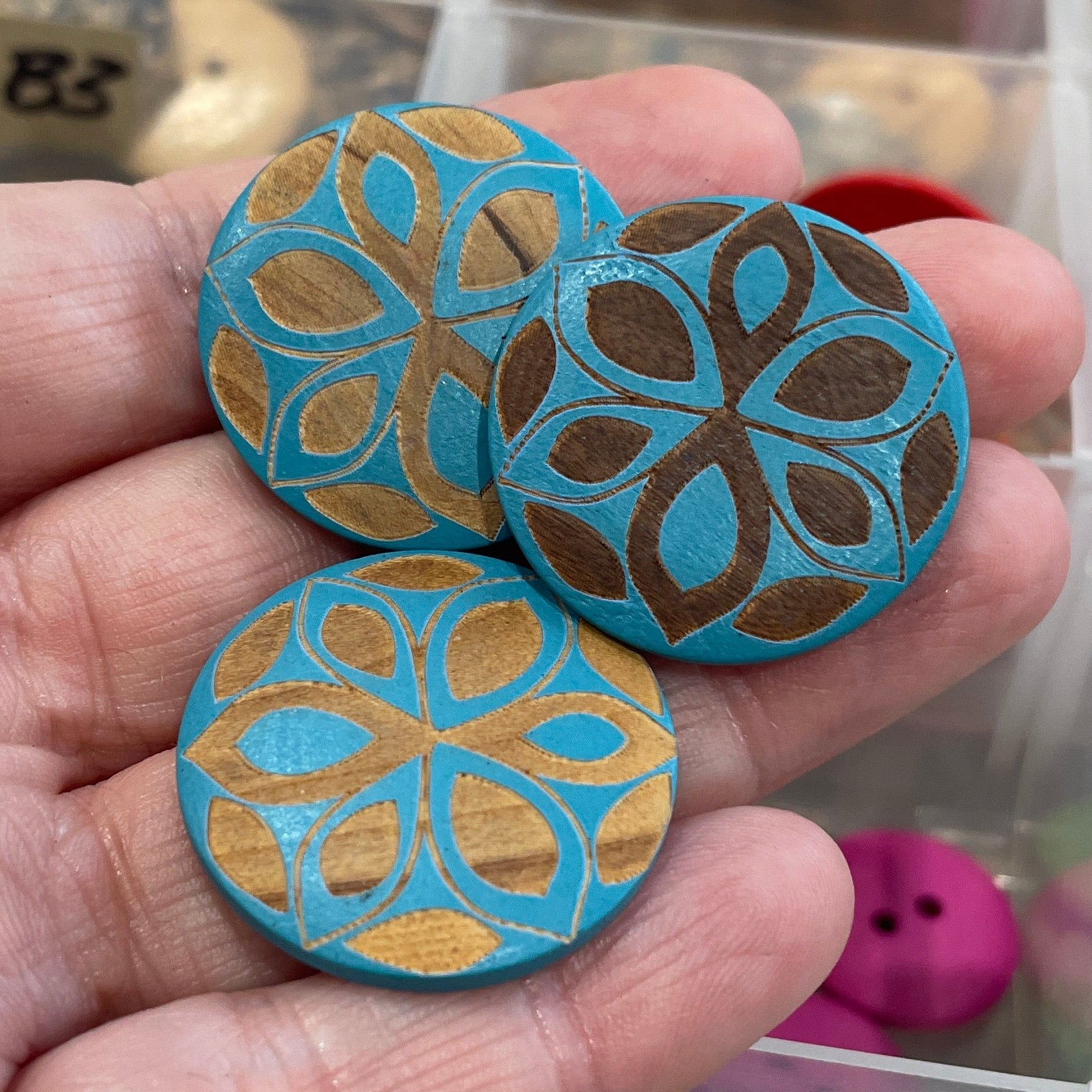 Hand holding decorative buttons with a patterned design in front of a container with more buttons.