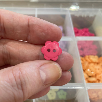 Hand holding a pink flower-shaped button with a blurred background of a storage container with buttons.