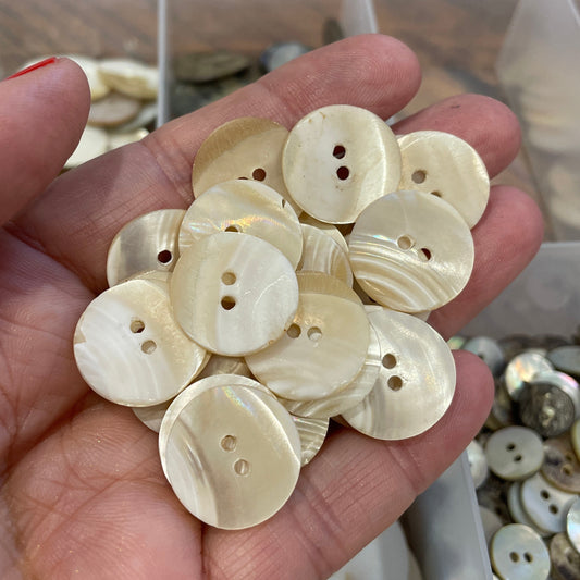 Hand holding a collection of cream-colored buttons with a blurred container of buttons in the background.