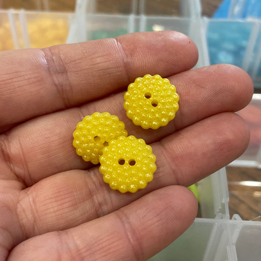 Three yellow button-like objects held in a hand with a blurred background of buttons and shelves.