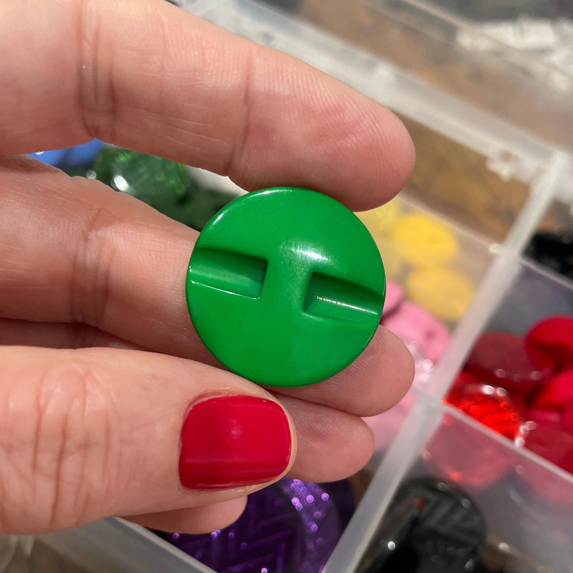 Hand holding a green plastic button with a letter 'H' in front of a container of colorful buttons.