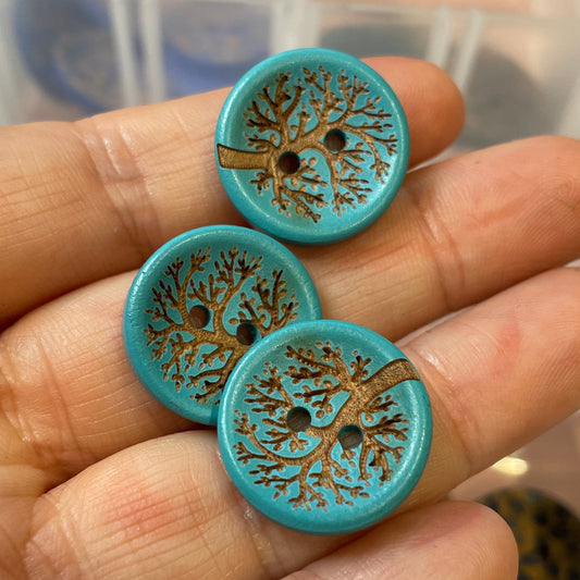 Three blue buttons with tree designs held by a hand, with a clear container of more buttons in the background.