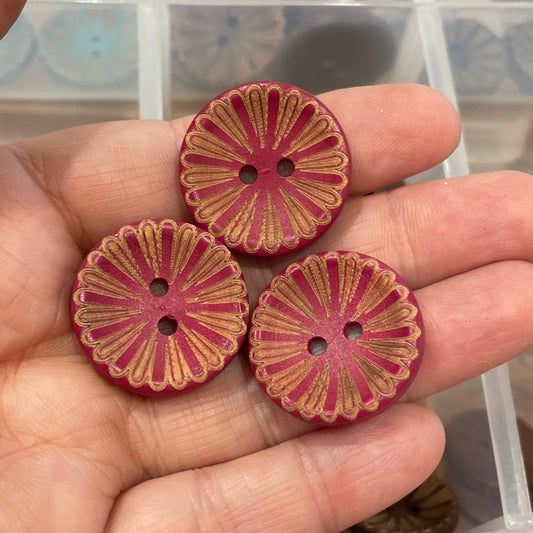 Three pink floral-patterned buttons held in a hand with a container of buttons in the background.