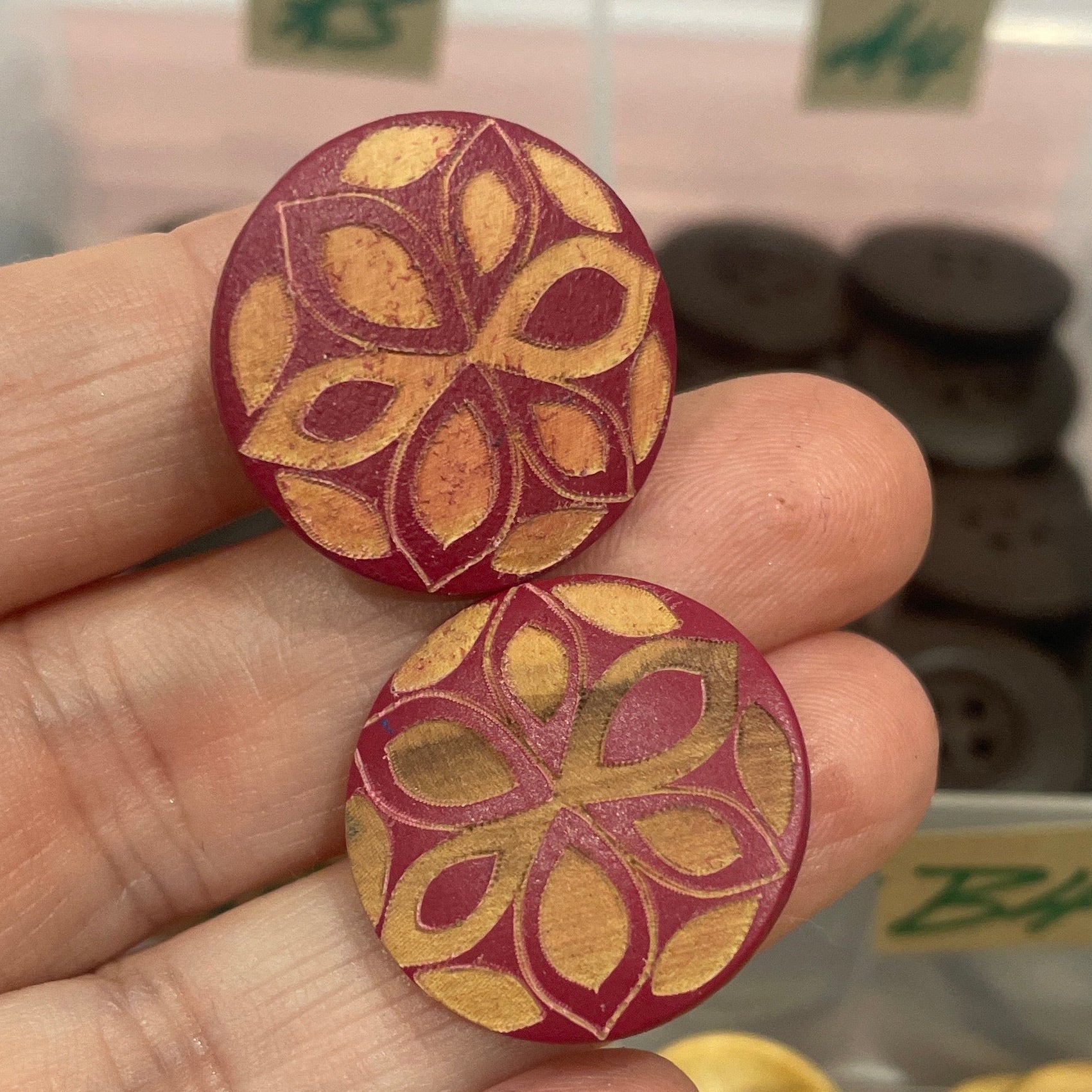 Two decorative buttons held between fingers with a container of buttons in the background.