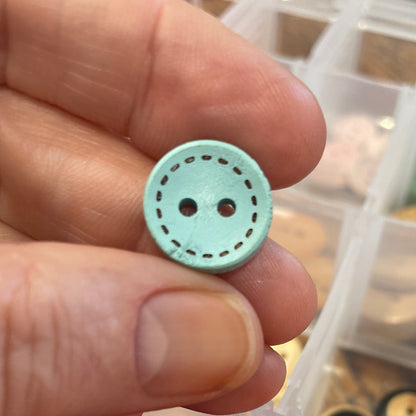 Hand holding a light blue button with a container of colorful buttons in the background.