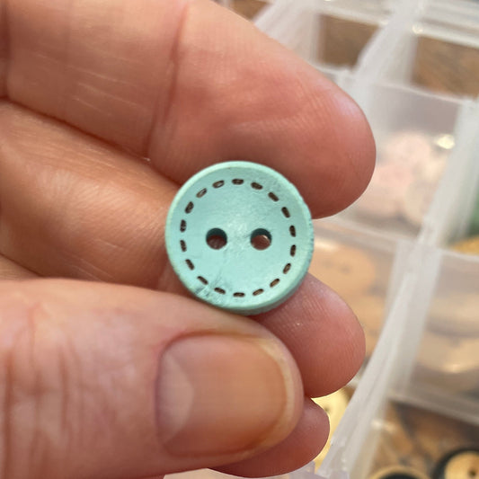 Hand holding a light blue button with a container of colorful buttons in the background.