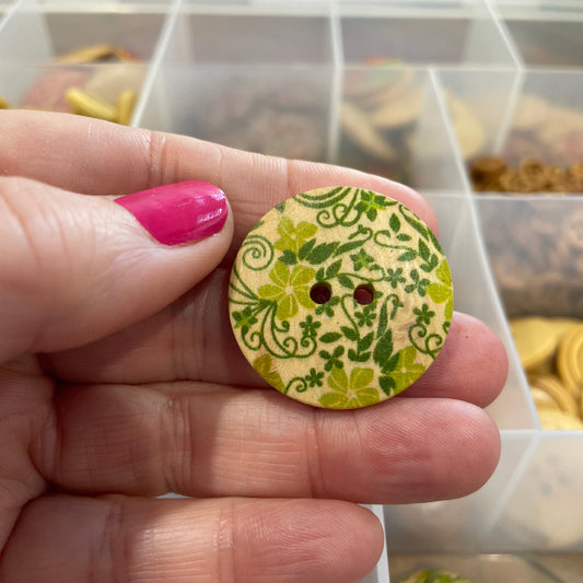 Hand holding a floral-patterned wooden button with a blurred background of more buttons.