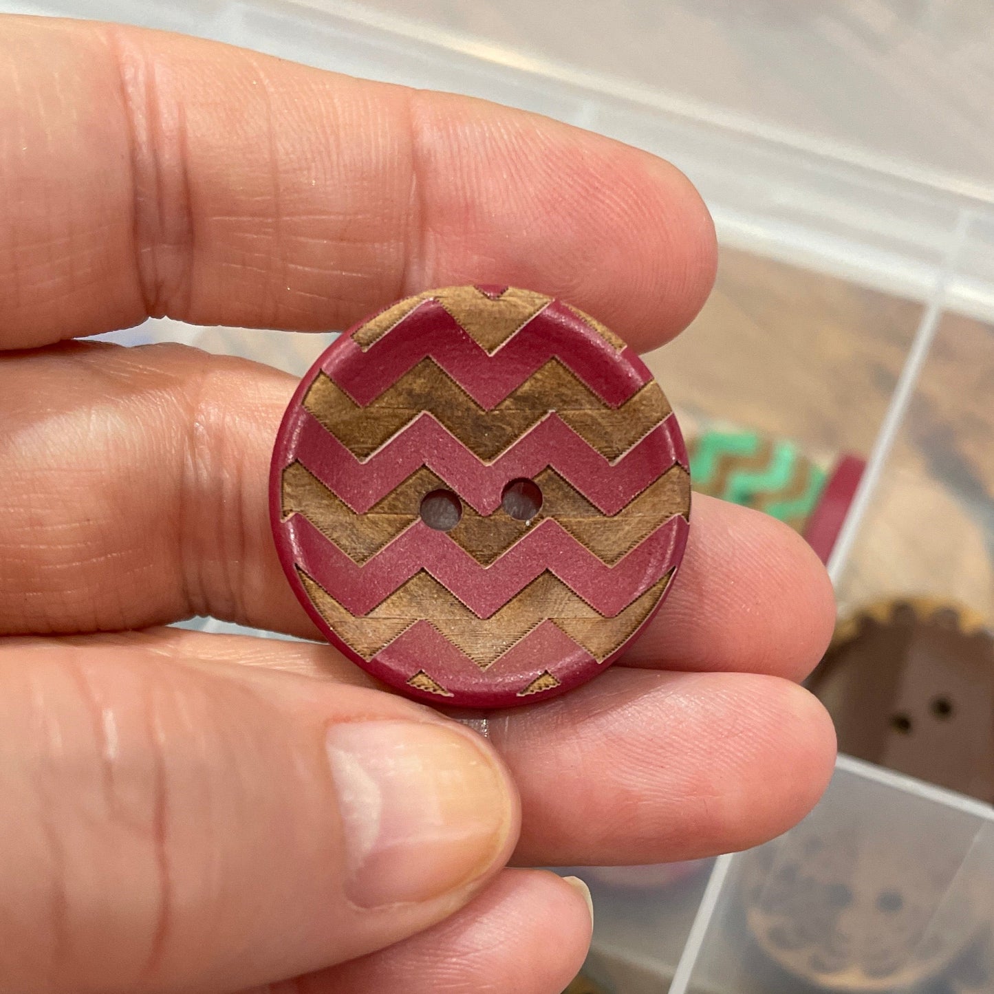 Hand holding a decorative button with a zigzag pattern, set against a background of various buttons in a container.