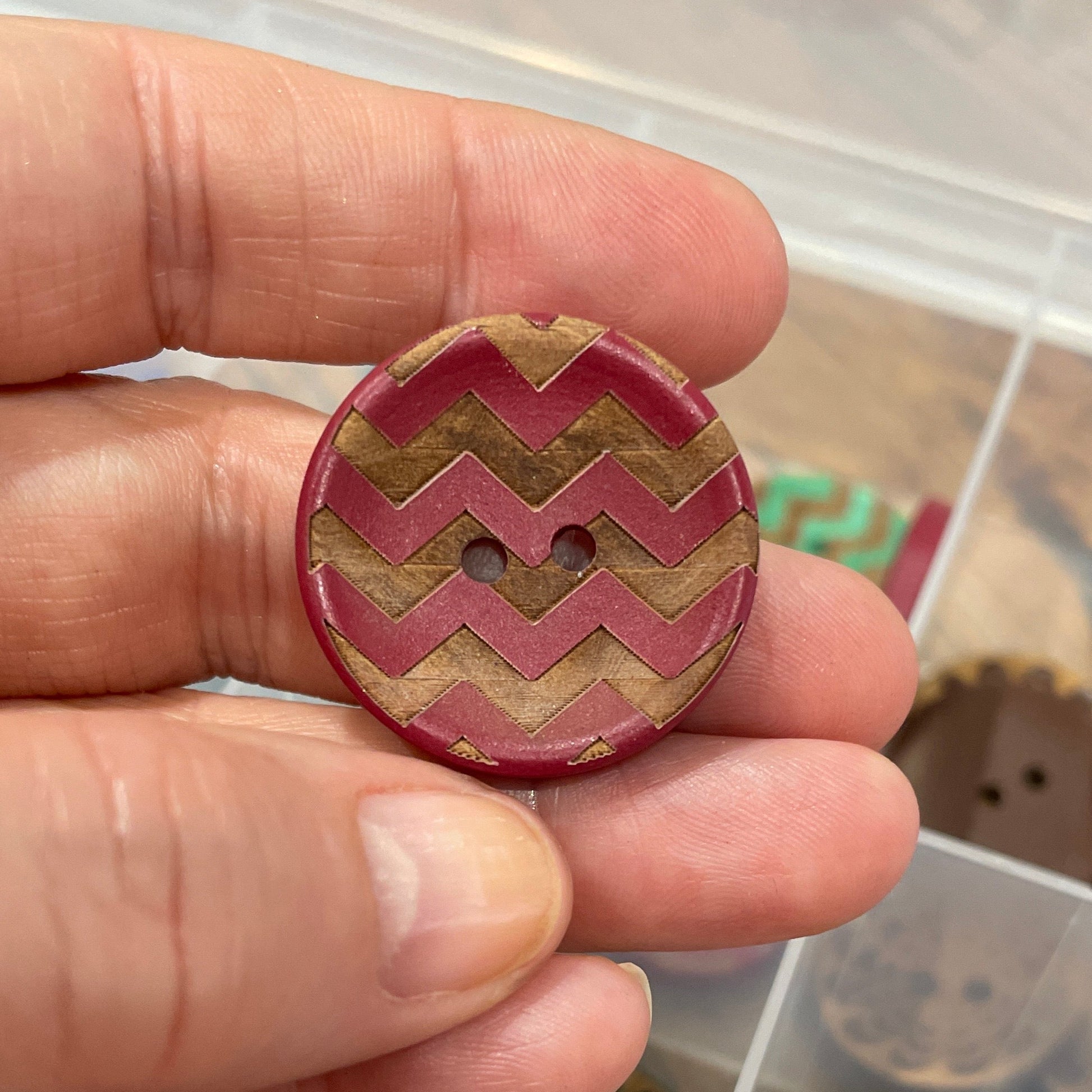 Hand holding a decorative button with a zigzag pattern, set against a background of various buttons in a container.