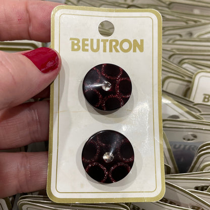 Pair of buttons on a Beutron branded card held by a hand with red nail polish.